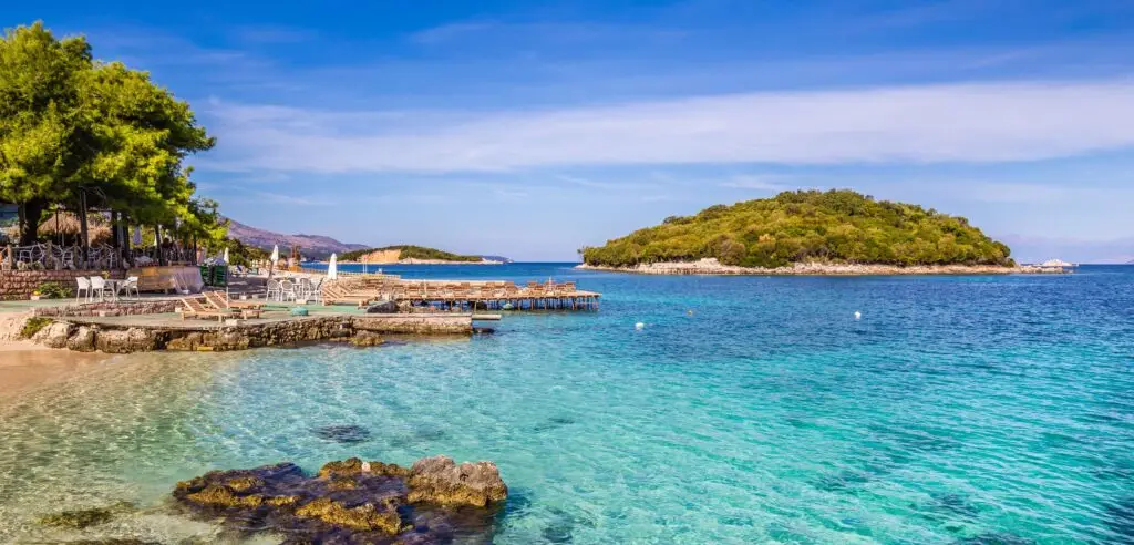 Plage de Vlorë en Albanie avec eau turquoise, pontons en bois et îlot boisé en arrière-plan