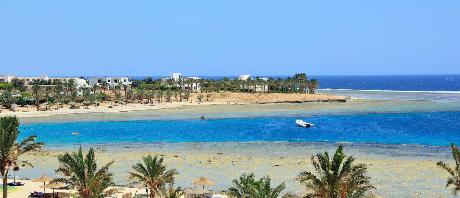 Lagon turquoise à Marsa Alam en mer Rouge avec récif corallien, plage bordée de palmiers et bateau de plongée au large